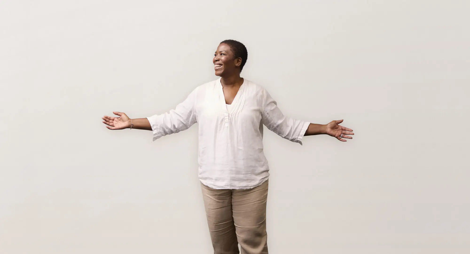 A person smiling with arms outstretched, wearing a white shirt and beige pants, standing against a plain white background.
