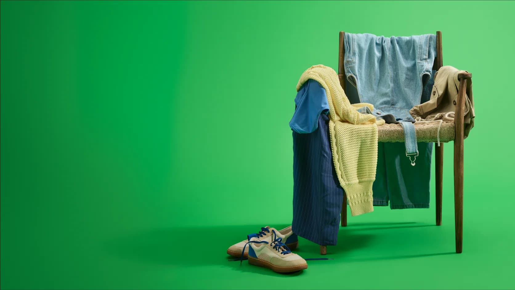 A wooden chair draped with various clothing items, including shirts and sweaters, against a solid green background. Shoes are placed on the floor.