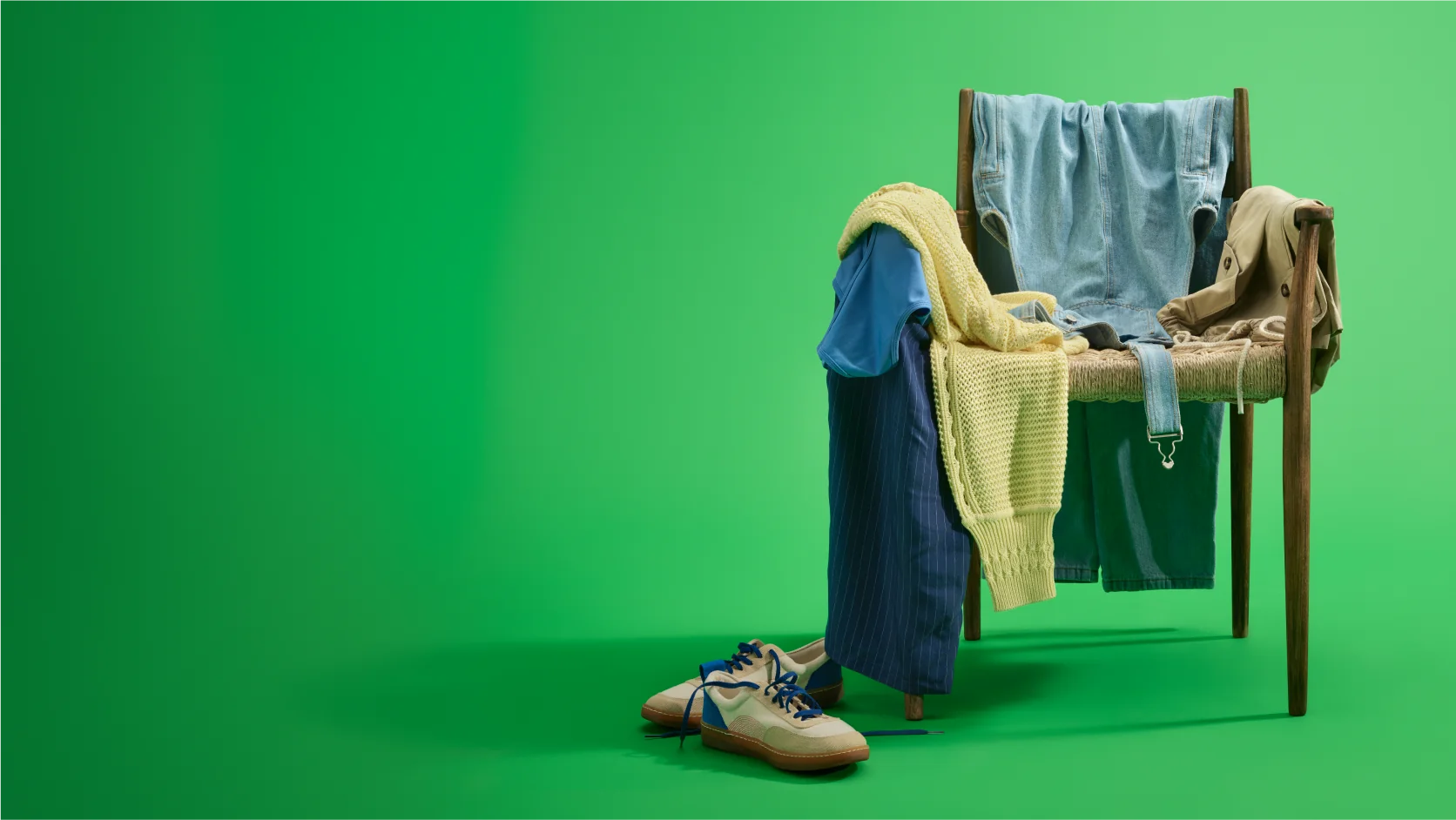 A wooden chair draped with various clothing items, including shirts and sweaters, against a solid green background. Shoes are placed on the floor.